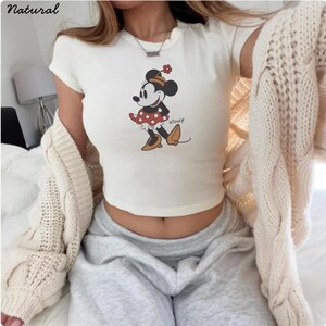May include: A cream-coloured crop top with a Minnie Mouse graphic. The cartoon character is wearing a red polka-dot dress, black shoes, and a matching hat. The top has short sleeves and a rounded neckline. The word "Disney" is printed below the graphic.