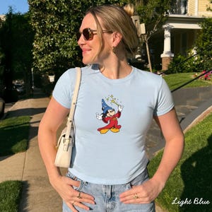 Może przedstawiać: Jasnoniebieska koszulka z nadrukiem kresk&oacute;wki Myszki Miki w stroju czarodzieja, trzymającego magiczną r&oacute;żdżkę z gwiazdami. Koszulka jest połączona z niebieskimi dżinsami. Tekst "Light Blue" jest widoczny w prawym dolnym rogu.