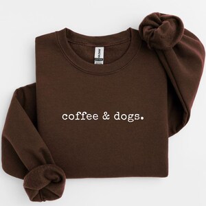 May include: Dark chocolate-colored sweatshirt with the text "coffee & dogs." in white. The long-sleeved crewneck top is folded neatly. A small, circular label in the upper right corner reads "DARK CHOCO."