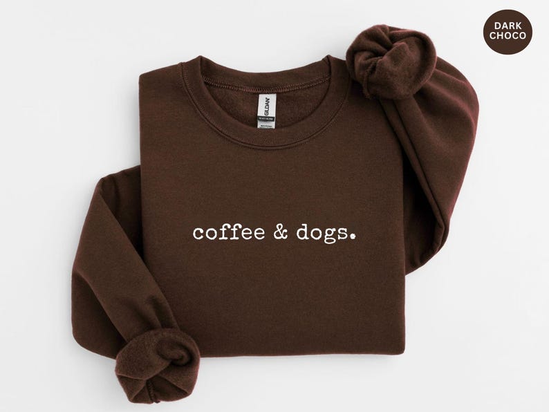 May include: Dark chocolate-colored sweatshirt with the text "coffee & dogs." in white. The long-sleeved crewneck top is folded neatly. A small, circular label in the upper right corner reads "DARK CHOCO."