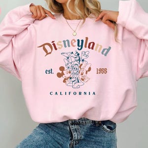 May include: Pink Disneyland sweatshirt with a colorful graphic featuring Mickey Mouse, Donald Duck, and other Disney characters. The text "Disneyland est. 1955 CALIFORNIA" is printed on the front.