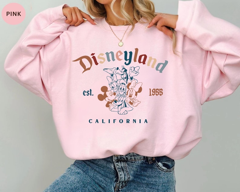 May include: Pink Disneyland sweatshirt with a colorful graphic featuring Mickey Mouse, Donald Duck, and other Disney characters. The text "Disneyland est. 1955 CALIFORNIA" is printed on the front.