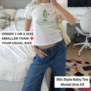 May include: White baby tee with a graphic of a leprechaun holding a beer and the text "DID SOMEONE SAY PADDY PARTY?". The shirt is paired with blue jeans. The text on the image says "ORDER 1 OR 2 SIZE SMALLER THAN YOUR USUAL SIZE" and "90s Style Baby Tee Model Size XS".
