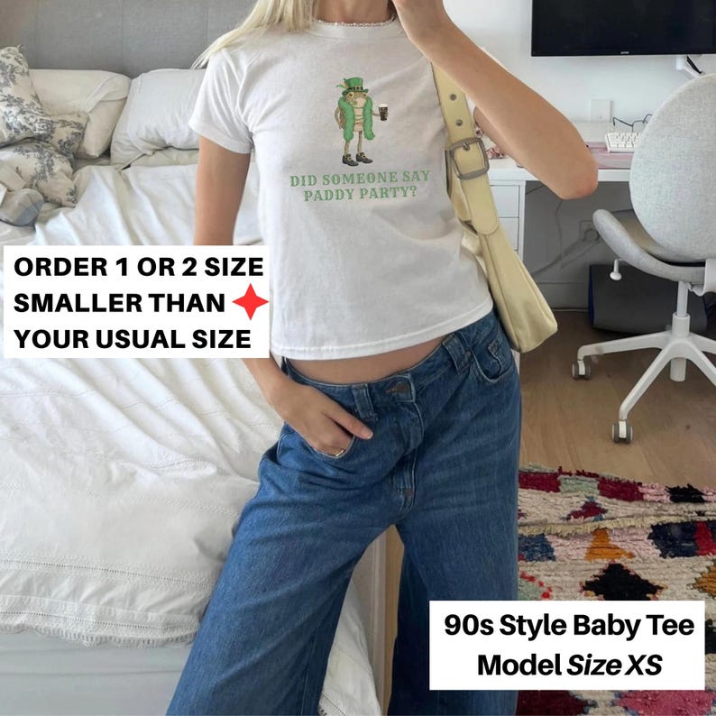 May include: White baby tee with a graphic of a leprechaun holding a beer and the text "DID SOMEONE SAY PADDY PARTY?". The shirt is paired with blue jeans. The text on the image says "ORDER 1 OR 2 SIZE SMALLER THAN YOUR USUAL SIZE" and "90s Style Baby Tee Model Size XS".