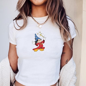 Może przedstawiać: Biały top z nadrukiem przedstawiającym Myszka Miki w stroju czarodzieja, trzymającego r&oacute;żdżkę z gwiazdami. Postać z kresk&oacute;wki ma na sobie niebieski kapelusz, czerwoną szatę i ż&oacute;łte buty. Top ma okrągły dekolt i kr&oacute;tkie rękawy.