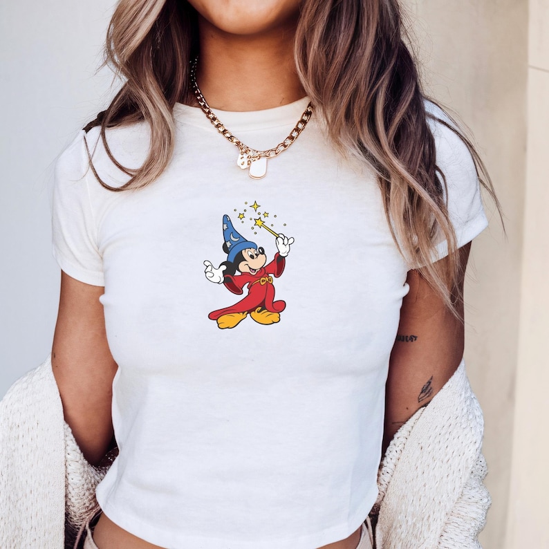 Może przedstawiać: Biały top z nadrukiem przedstawiającym Myszka Miki w stroju czarodzieja, trzymającego r&oacute;żdżkę z gwiazdami. Postać z kresk&oacute;wki ma na sobie niebieski kapelusz, czerwoną szatę i ż&oacute;łte buty. Top ma okrągły dekolt i kr&oacute;tkie rękawy.