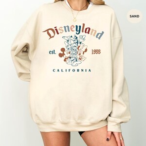 May include: Sand-colored Disneyland sweatshirt with a vintage design. The graphic features Mickey Mouse, Donald Duck, Goofy, and Minnie Mouse. The text includes "Disneyland," "est. 1955," and "California" in various colors.