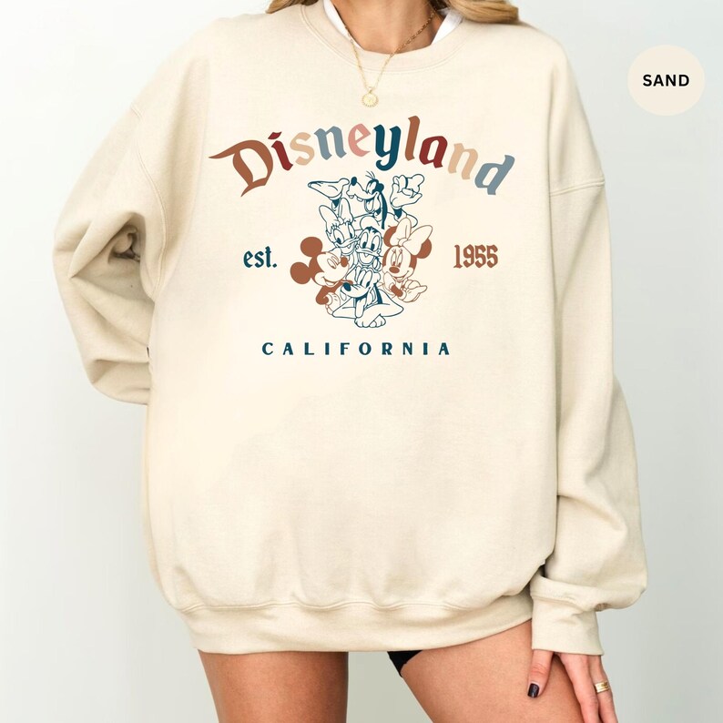 May include: Sand-colored Disneyland sweatshirt with a vintage design. The graphic features Mickey Mouse, Donald Duck, Goofy, and Minnie Mouse. The text includes "Disneyland," "est. 1955," and "California" in various colors.