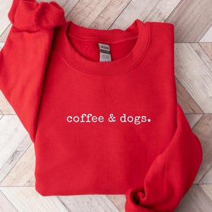 May include: A red sweatshirt with the text "coffee & dogs" in white. The crew neck sweatshirt is made of a soft material and is displayed on a wooden surface. A red circle with the word "RED" is in the upper right corner.