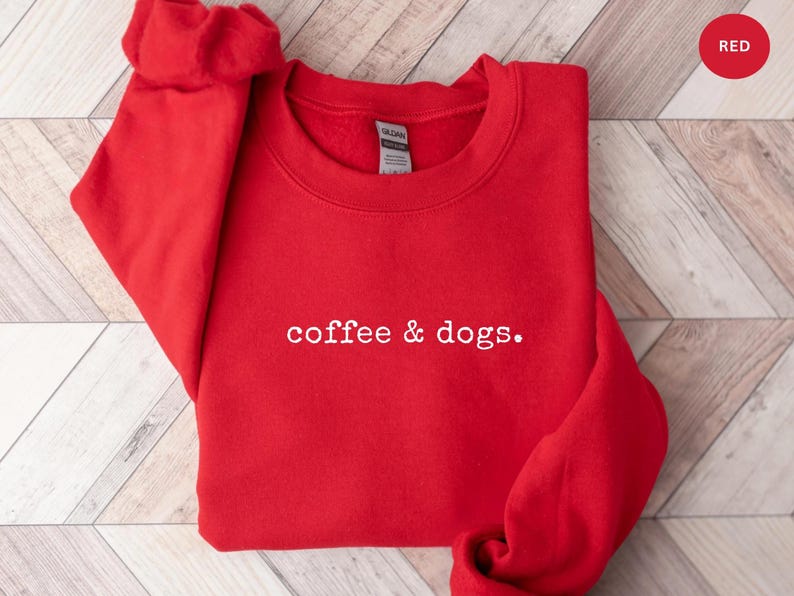 May include: A red sweatshirt with the text "coffee & dogs" in white. The crew neck sweatshirt is made of a soft material and is displayed on a wooden surface. A red circle with the word "RED" is in the upper right corner.
