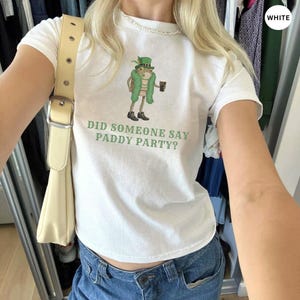 May include: White t-shirt with a green graphic of a leprechaun holding a beer and the text "DID SOMEONE SAY PADDY PARTY?". The shirt is paired with blue jeans and a cream-colored purse.