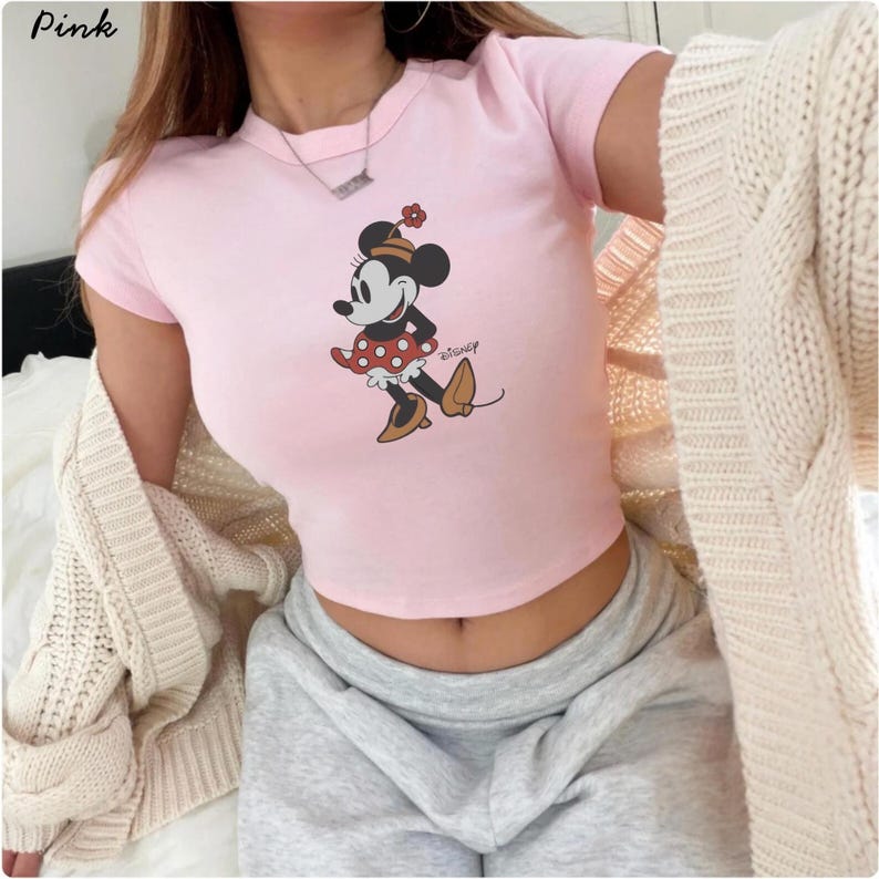 May include: Pale pink crop top with a Minnie Mouse design. The cartoon character is shown in a classic pose, wearing a red polka-dot dress and brown shoes. The top has short sleeves and a round neckline. The word "Disney" is printed on the shirt.