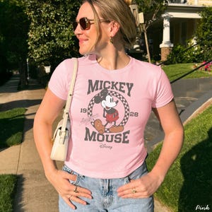Pu&ograve; includere: T-shirt rosa chiaro con grafica vintage di Topolino. Il design include le parole "Mickey Mouse" e gli anni "19" e "28" attorno a un'immagine a cartoni animati di Topolino. La maglietta ha maniche corte e girocollo.