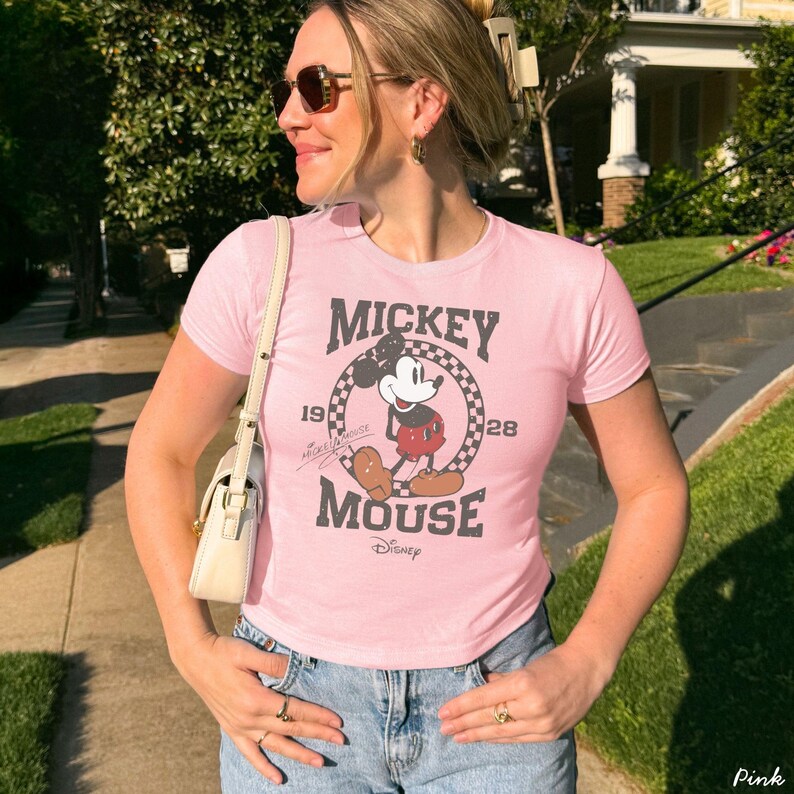 Pu&ograve; includere: T-shirt rosa chiaro con grafica vintage di Topolino. Il design include le parole "Mickey Mouse" e gli anni "19" e "28" attorno a un'immagine a cartoni animati di Topolino. La maglietta ha maniche corte e girocollo.