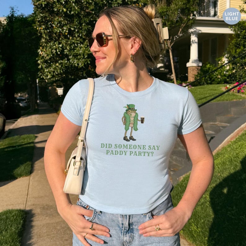 May include: Light blue t-shirt with a cartoon leprechaun holding a mug of beer. The shirt has the text "DID SOMEONE SAY PADDY PARTY?" printed below the graphic. The model is wearing sunglasses and jeans.