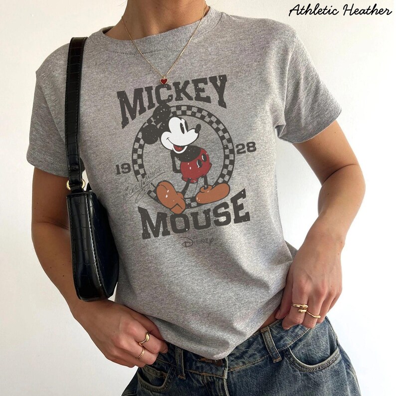 Pu&ograve; includere: T-shirt grigio athletic heather con grafica di Mickey Mouse invecchiata. Il design include le parole "Mickey Mouse" e l'anno "1928". La maglietta ha girocollo e maniche corte.