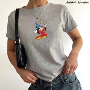 Może przedstawiać: Szara koszulka z grafiką Myszki Miki jako czarodzieja, trzymającego r&oacute;żdżkę z gwiazdami. Tekst "Athletic Heather" jest wydrukowany w prawym g&oacute;rnym rogu. Koszulka jest połączona z niebieskimi dżinsami i czarną torebką.