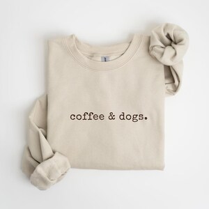 May include: Sand-colored sweatshirt with the embroidered text "coffee & dogs." The long-sleeved top is folded, with the sleeves rolled up. The sweatshirt is made of a soft material and is suitable for casual wear.