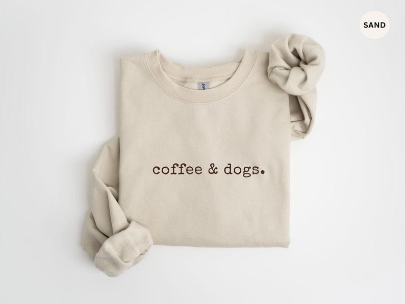 May include: Sand-colored sweatshirt with the embroidered text "coffee & dogs." The long-sleeved top is folded, with the sleeves rolled up. The sweatshirt is made of a soft material and is suitable for casual wear.