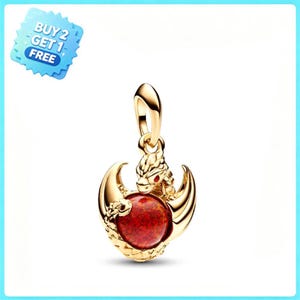 Puede incluir: Un colgante de tono dorado que presenta una figura de dragón esculpida con un pequeño ojo rojo, que sujeta una piedra roja grande, redonda y texturizada en sus garras. El cuerpo del dragón se curva alrededor de la piedra y tiene una argolla.
