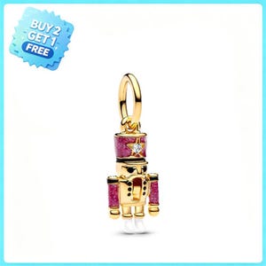 May include: A gold-toned nutcracker charm featuring a glittery magenta hat and sleeves, a small clear stone set in a star on the hat, and white painted feet. The charm includes a circular bail at the top for attachment to a bracelet or necklace chain.
