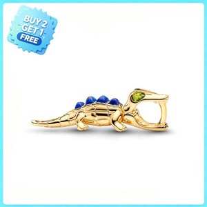 May include: A gold-toned crocodile charm featuring a series of blue stone accents along its back and a single green stone set in its head. The charm is designed with an open mouth loop for attachment, and a blue starburst graphic with the text BUY 2 GET 1 FREE.