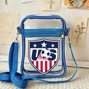 May include: A transparent crossbody bag with a blue zip and strap. The bag displays a shield emblem with "US", a football, and red and white stripes, representing the United States.