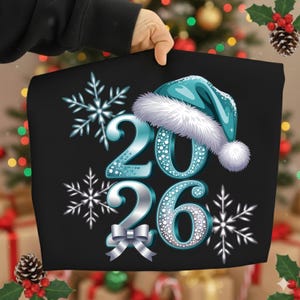Puede incluir: Gráfico negro con los números "2026" en verde azulado con detalles de puntos blancos, un gorro de Papá Noel verde azulado con ribete blanco, copos de nieve y un lazo plateado. Ideal para ropa festiva.