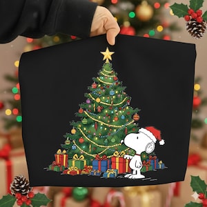 Puede incluir: Bolso tote negro con temática navideña con un árbol de Navidad decorado y Snoopy con un gorro de Papá Noel. El árbol está adornado con adornos y luces, y rodeado de regalos envueltos.