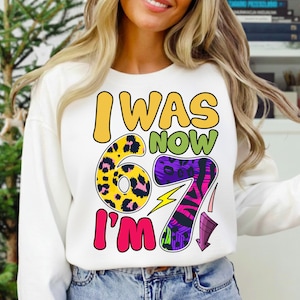 Peut inclure: Sweat-shirt blanc avec le texte "I WAS NOW 67 I'M". Le chiffre 6 est jaune avec un motif léopard, et le chiffre 7 est violet avec un motif zébré. Un éclair jaune est entre les chiffres.