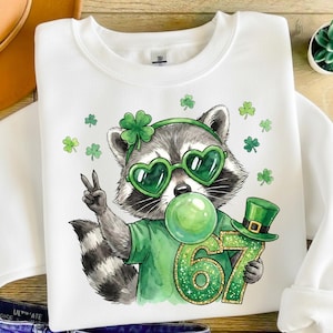 Puede incluir: Sudadera blanca con un mapache que lleva gafas de sol verdes en forma de corazón, una diadema verde con un trébol y una camiseta verde. El mapache está soplando una burbuja verde y sosteniendo un sombrero verde y el número 67. Tréboles verdes dispersos.