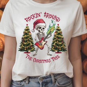 Könnte beinhalten: Weißes T-Shirt mit einem Skelett, das Gitarre spielt, einen Weihnachtsmannhut trägt und von Weihnachtsbäumen flankiert wird. Der Text lautet "Rockin' Around The Christmas Tree" in roter Schrift.
