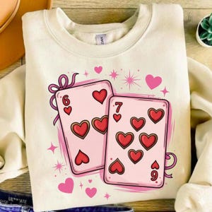 Puede incluir: Sudadera de color crema con un gráfico de dos cartas de juego con corazones. Las cartas son rosas con símbolos de corazón rojos y los números 6 y 7. Corazones y estrellas rosas están dispersos alrededor de las cartas.
