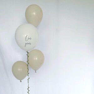 May include: A cluster of four balloons in cream and white. The largest balloon is white with the words "One year" in a delicate script. A green vine with small leaves adds a natural touch to the arrangement. Perfect for a first birthday celebration.
