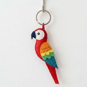 May include: A handmade felt parrot keychain. The parrot is primarily red with a white head, a blue beak, and colorful wings in orange, yellow, green, and blue. The keychain is attached to a silver metal ring.
