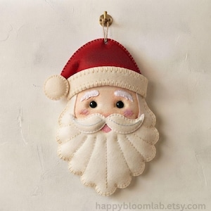 Op de afbeelding: Een vilten kerstman-ornament met een rode muts met witte pompon, een crèmekleurige baard en roze wangen. Het ornament heeft zwarte kraaloogjes, witte wenkbrauwen en een gestikte snor. Het hangt aan een klein gouden haakje aan de bovenkant.