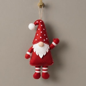 May include: A felt gnome ornament featuring a red hat with white polka dots and a white pom-pom tip. The gnome has a white beard, a beige nose, and a red body with white-striped arms and legs. It hangs from a thin string attached to a gold hook.