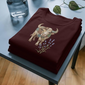Könnte beinhalten: Ein Stapel von zwei weinroten T-Shirts. Jedes Shirt zeigt eine detaillierte Illustration eines Stiers mit Patchwork-Design und Lavendelzweigen. Der Stier hat ein Blumenmuster und ist in Braun-, Beige- und Blautönen gehalten.