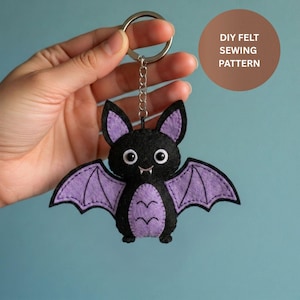 Puede incluir: Un llavero de fieltro negro con forma de murciélago con alas y detalles morados. El murciélago tiene ojos blancos, una boca pequeña y un llavero plateado. La imagen incluye el texto "DIY FELT SEWING PATTERN" en un círculo marrón.