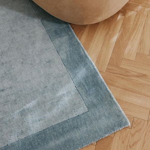 May include: A close-up of a blue rug with a lighter blue border. The rug is on a wooden floor with a beige beanbag chair in the background. The rug has a soft, textured appearance.