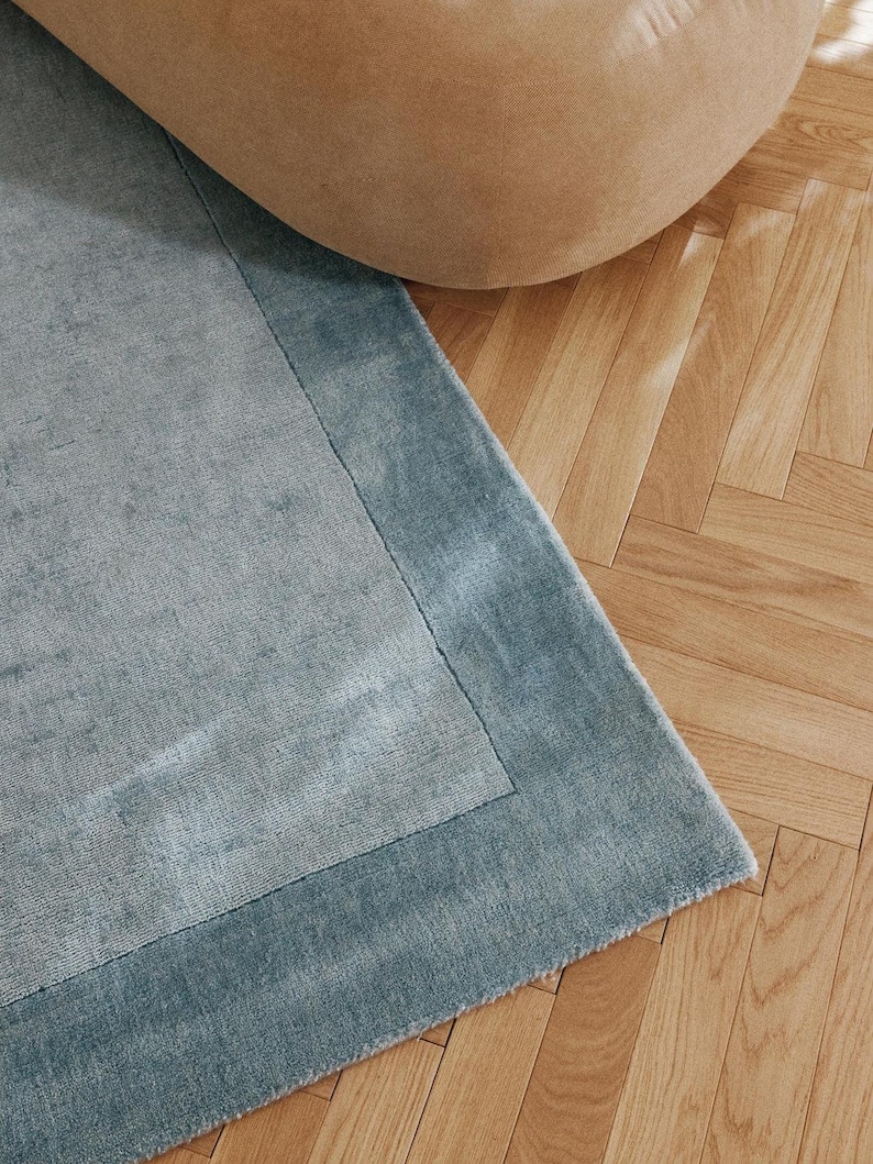 May include: A close-up of a blue rug with a lighter blue border. The rug is on a wooden floor with a beige beanbag chair in the background. The rug has a soft, textured appearance.