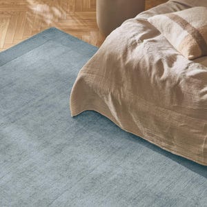 May include: A light blue rectangular rug with a textured surface and a darker blue border. The rug is placed on a wooden floor, partially under a bed with beige bedding and a striped pillow. A small side table is visible.
