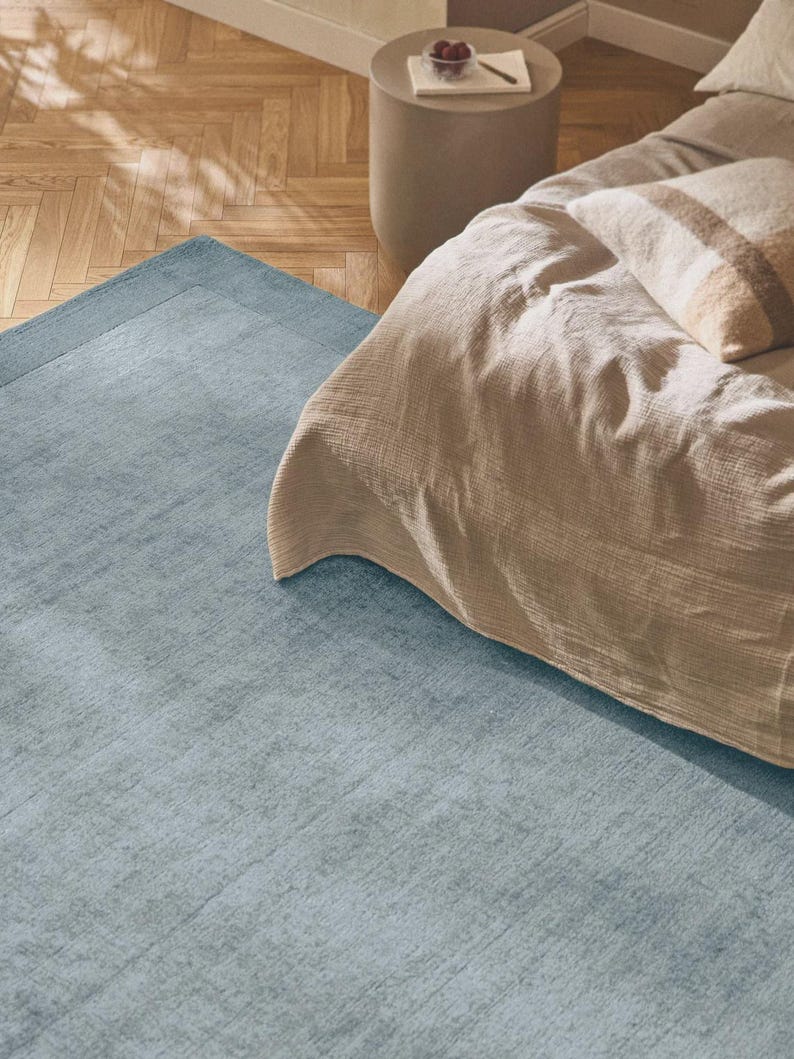 May include: A light blue rectangular rug with a textured surface and a darker blue border. The rug is placed on a wooden floor, partially under a bed with beige bedding and a striped pillow. A small side table is visible.