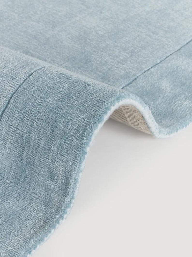 May include: Close-up of a light blue rug with a soft, textured surface. The rug's edge is slightly curled, revealing a beige backing. The rug has a subtle sheen, suggesting a plush material. The overall aesthetic is clean and modern.