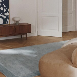 May include: A light blue rectangular rug with a darker border is placed on a herringbone wood floor. A tan, rounded sofa is partially visible on the right. A wooden cabinet with a lamp and artwork is in the background.