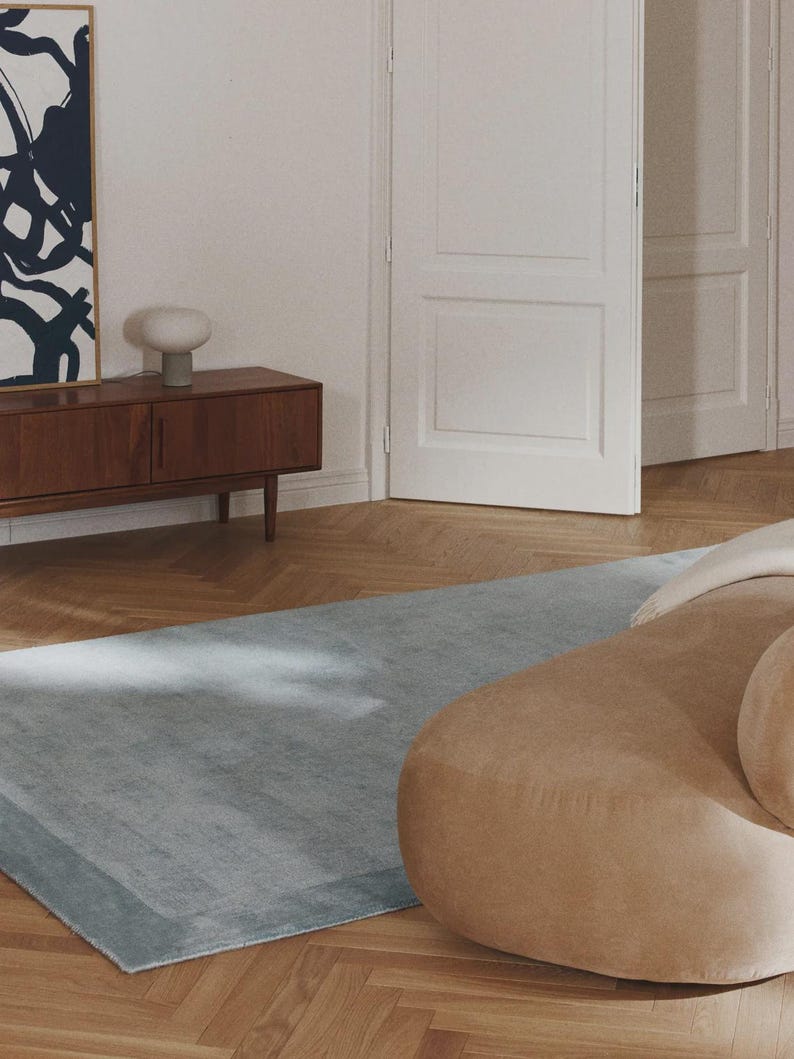 May include: A light blue rectangular rug with a darker border is placed on a herringbone wood floor. A tan, rounded sofa is partially visible on the right. A wooden cabinet with a lamp and artwork is in the background.