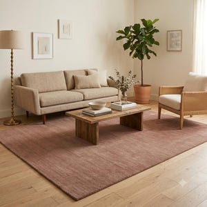 May include: A large rectangular area rug in a muted dusty rose color covering a light wood floor. The rug is positioned beneath a beige sofa, a wooden coffee table, and a cane-backed armchair, providing a soft, textured foundation for the room.