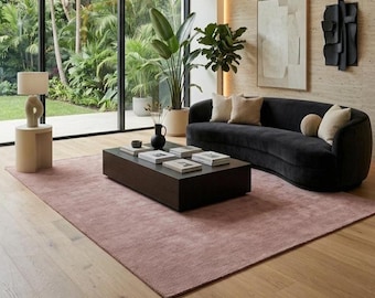 Op de afbeelding: Een woonkamer met een gebogen zwarte bank met beige kussens, een rechthoekige salontafel van donker hout met boeken, een roze vloerkleed, een beige cilindrische bijzettafel met een lamp en grote ramen die uitkijken op een groene tuin.
