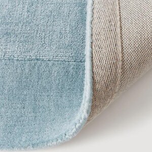 May include: Close-up of a light blue rug with a soft, textured surface. The rug has a light blue border and a beige backing with a herringbone pattern. The rug is partially rolled up, revealing the backing.