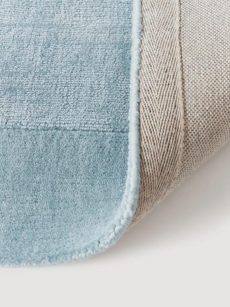 May include: Close-up of a light blue rug with a soft, textured surface. The rug has a light blue border and a beige backing with a herringbone pattern. The rug is partially rolled up, revealing the backing.